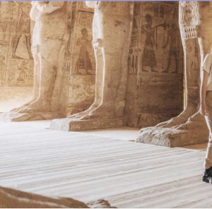 Inside an ancient Egyptian temple, massive stone statues' legs and bases set on wide stone floors with hieroglyphic carvings on the walls